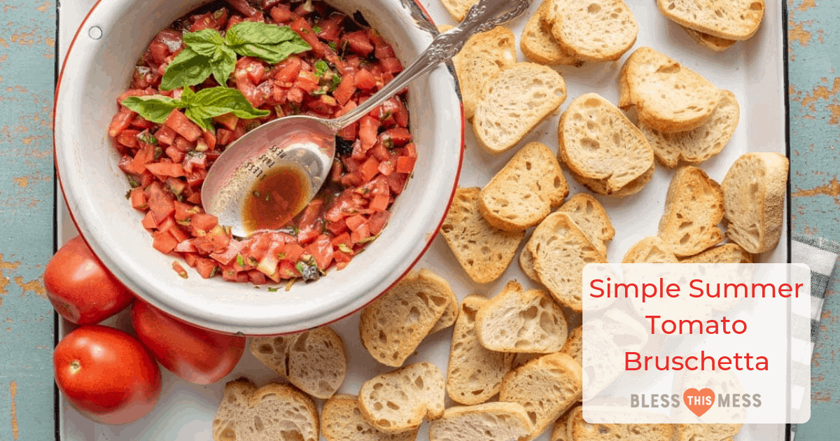 small slices of bread on a white sheet pan with red tomatoes and a white bowl of chopped tomatoes with green basil for summer tomato bruschetta and a silver spoon.