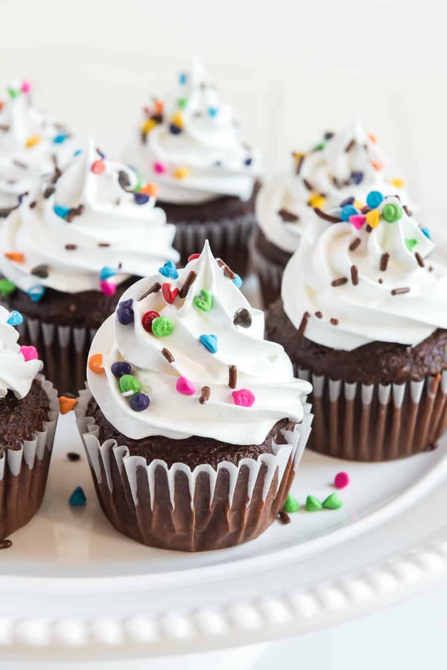 close up image of chocolate cupcakes with white frosting and multi-colored sprinkles all on a white plate.