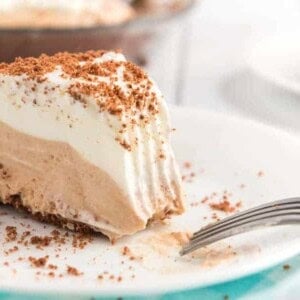 A slice of creamy chocolate peanut butter pie with layers of decadent chocolate and vanilla filling topped with a layer of whipped cream and sprinkled with crumbled chocolate on a white plate. A fork rests beside the slice, and another pie is blurred in the background.