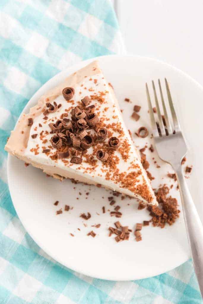 A slice of creamy dessert topped with chocolate shavings and fluffy icing sits on a white plate. A fork rests beside it, and a checkered blue and white cloth is partially visible in the background.