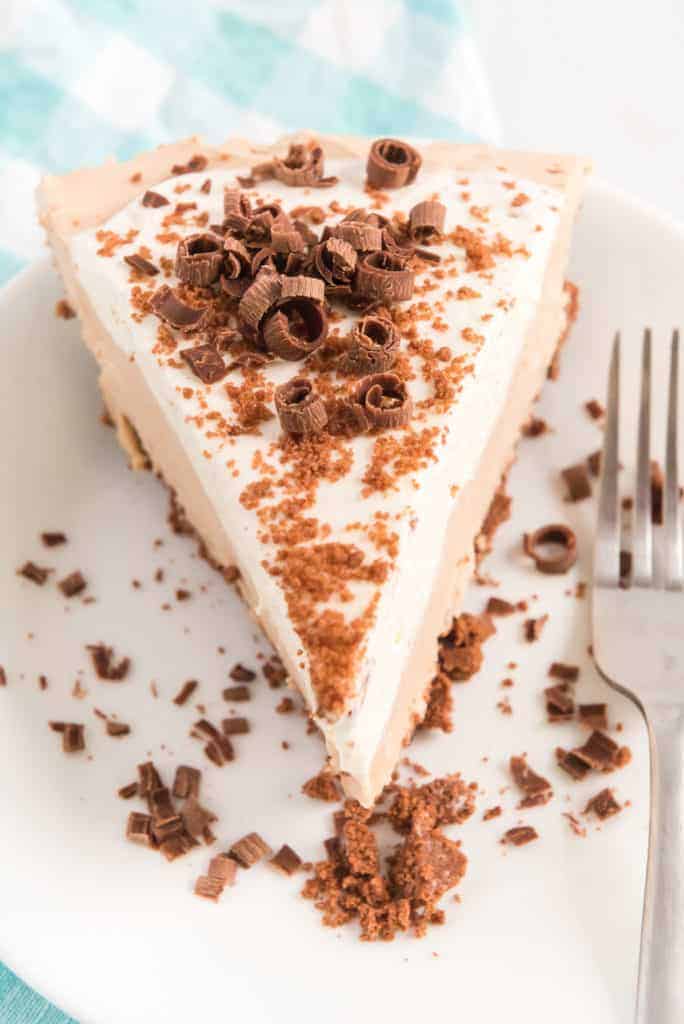 A slice of creamy pie with fluffy icing topped with shaved chocolate curls sits on a white plate. Crumbled chocolate bits are sprinkled on top and around the plate. A fork is placed on the plates edge, and a checkered cloth is partially visible in the background.