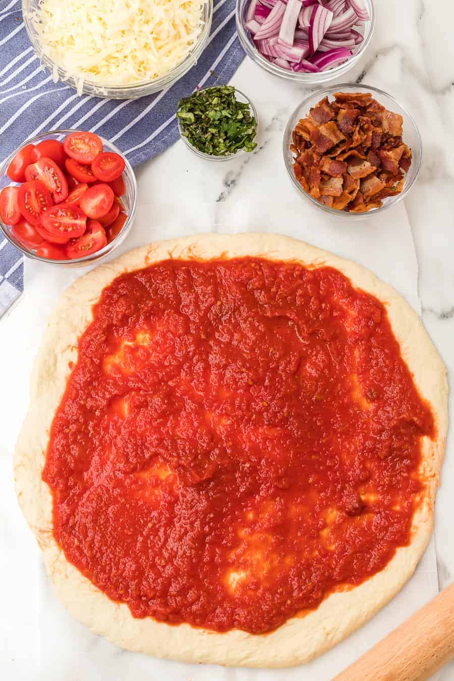 a raw pizza dough with red sauce and toppings for the a raw a whole herbed tomato and bacon pizza on parchment.