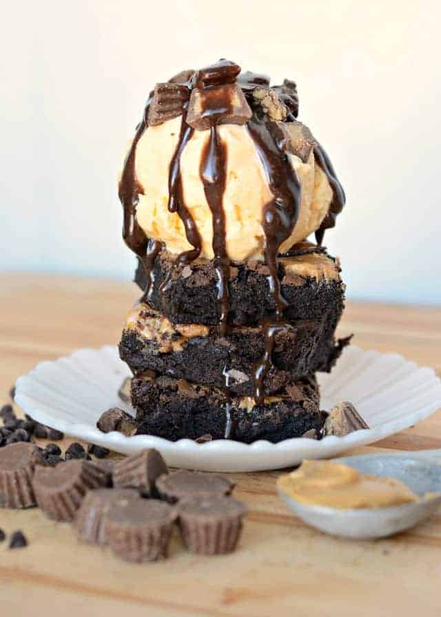 side view of brownies topped with ice cream, fudge sauce, and chopped peanut butter cups on a white plate