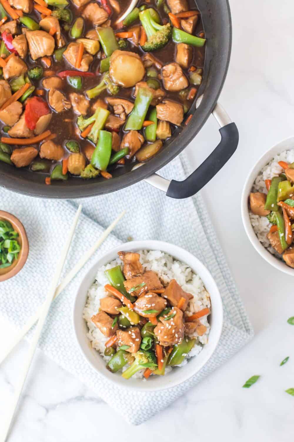 stir fried sticky saucy chicken and veggies in a pan and then the served on the side over rice in a white bowl.