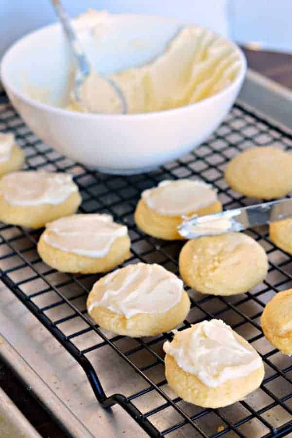 Baked melt away cookies on a black wire cooling rack being iced with the bowl of icing in the background. 