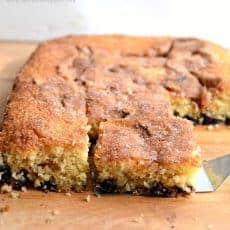 Close-up of a freshly baked cinnamon cake on a wooden surface. A spatula lifts one square piece from the cake, revealing a moist interior with a layer of berries at the bottom. The top is sprinkled with sugar, giving it a golden-brown crust.