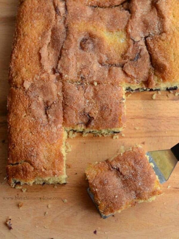 A cinnamon coffee cake with a golden-brown crust, reminiscent of a delectable blueberry boy bait, rests on a wooden surface. A square piece has been cut and is being lifted with a spatula, revealing its moist texture and sugar-cinnamon topping.