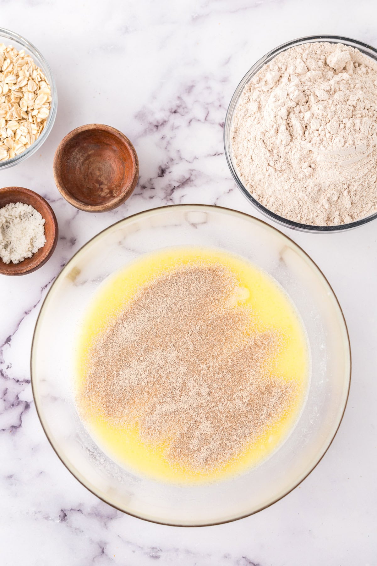 A bowl with melted butter and cinnamon sits on a marble countertop, surrounded by ingredients: a bowl of flour, oats, sugar, and an empty wooden bowl—perfect essentials for crafting whole wheat hamburger buns.