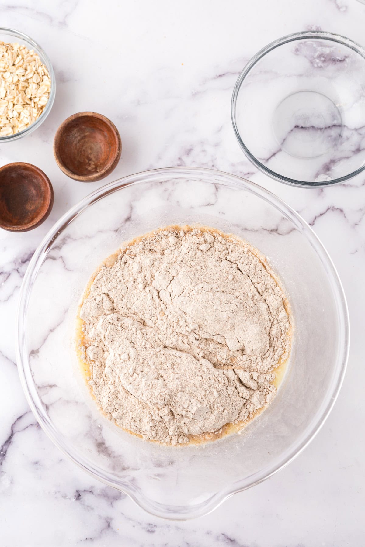 A glass mixing bowl brimming with flour and dry ingredients sits on a marble countertop, ready to craft homemade whole wheat hamburger buns. Nearby, small wooden bowls accompany a bowl of oats, while an empty glass bowl awaits its culinary duty.