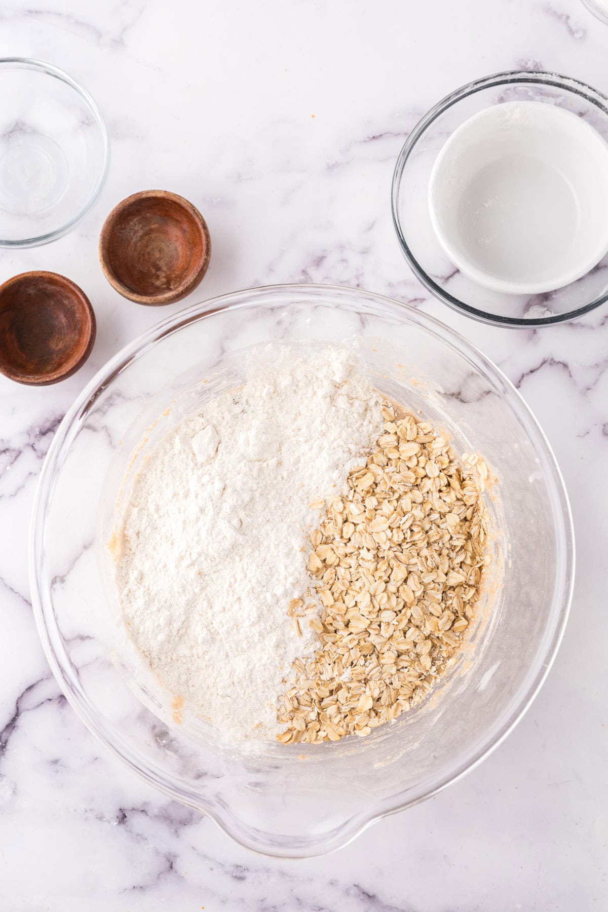 A glass mixing bowl containing flour and oats sits on a marble countertop, ready to be transformed into whole wheat hamburger buns. Surrounding it are small wooden bowls and empty glass bowls, all eager to join the culinary symphony of baking preparation.