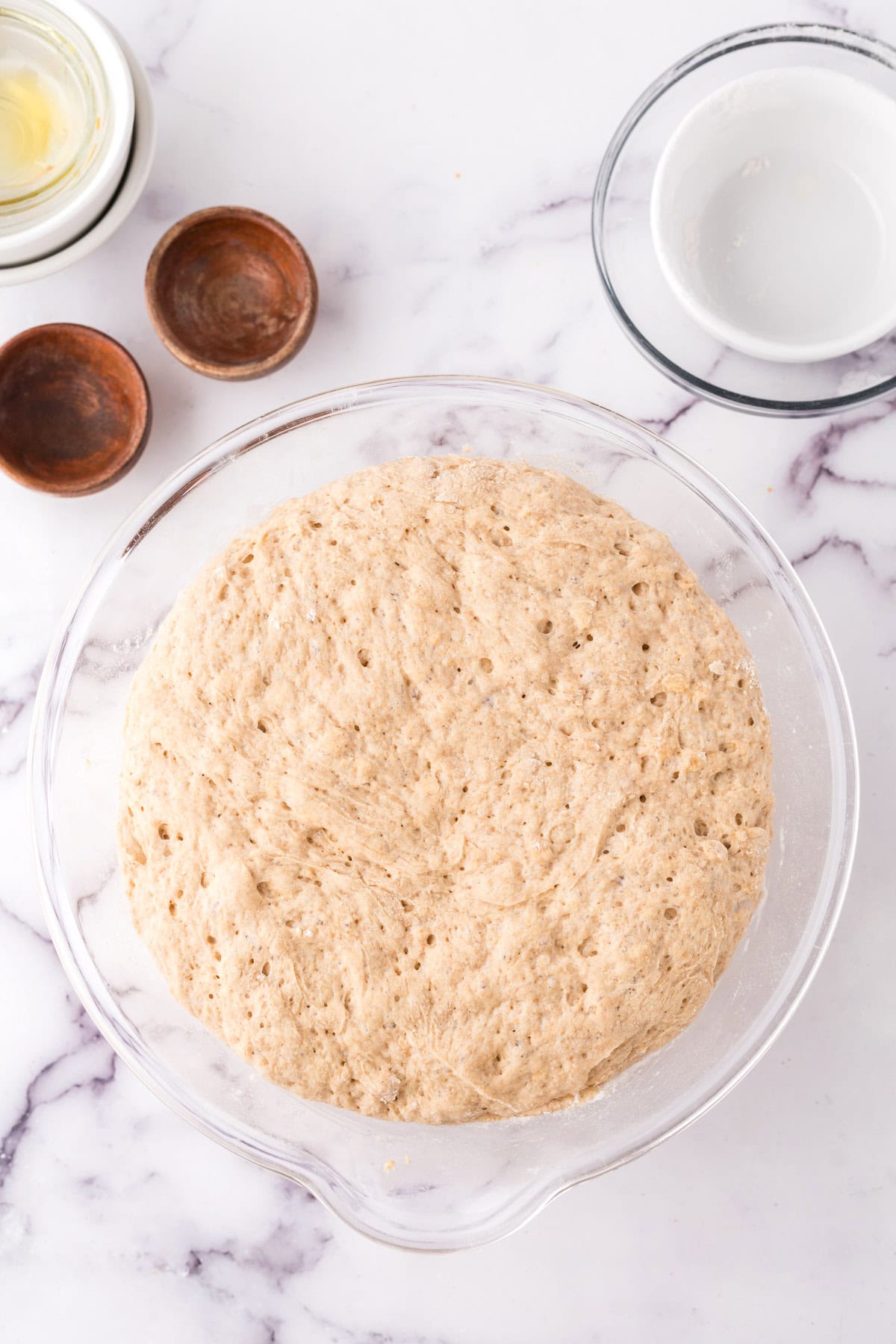 A clear glass bowl filled with freshly risen dough for whole wheat hamburger buns sits on a marble countertop. Surrounding it are small wooden bowls and two empty glass bowls, suggesting a baking preparation scene.