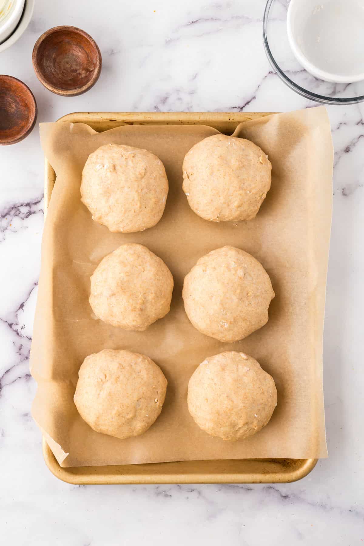 Six round whole wheat hamburger buns are arranged on a parchment-lined baking sheet. The sheet rests on a marble countertop, with small bowls and a plate partially visible in the background.