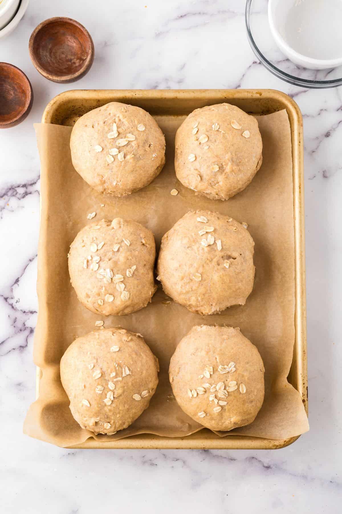 Six freshly baked whole wheat hamburger buns topped with oats are arranged on a parchment-lined baking sheet. The buns are golden-brown and sit on a marble countertop. Nearby, there are small brown bowls and a white mug.