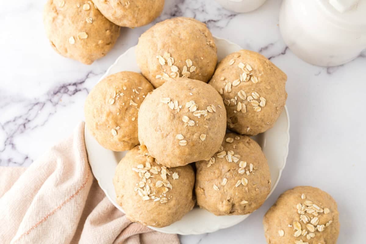 A plate of freshly baked oat rolls topped with oats rests on a marble surface, resembling whole wheat hamburger buns. One roll sits beside the plate. A folded cloth is on the left, and the edge of a milk jug is visible in the background.