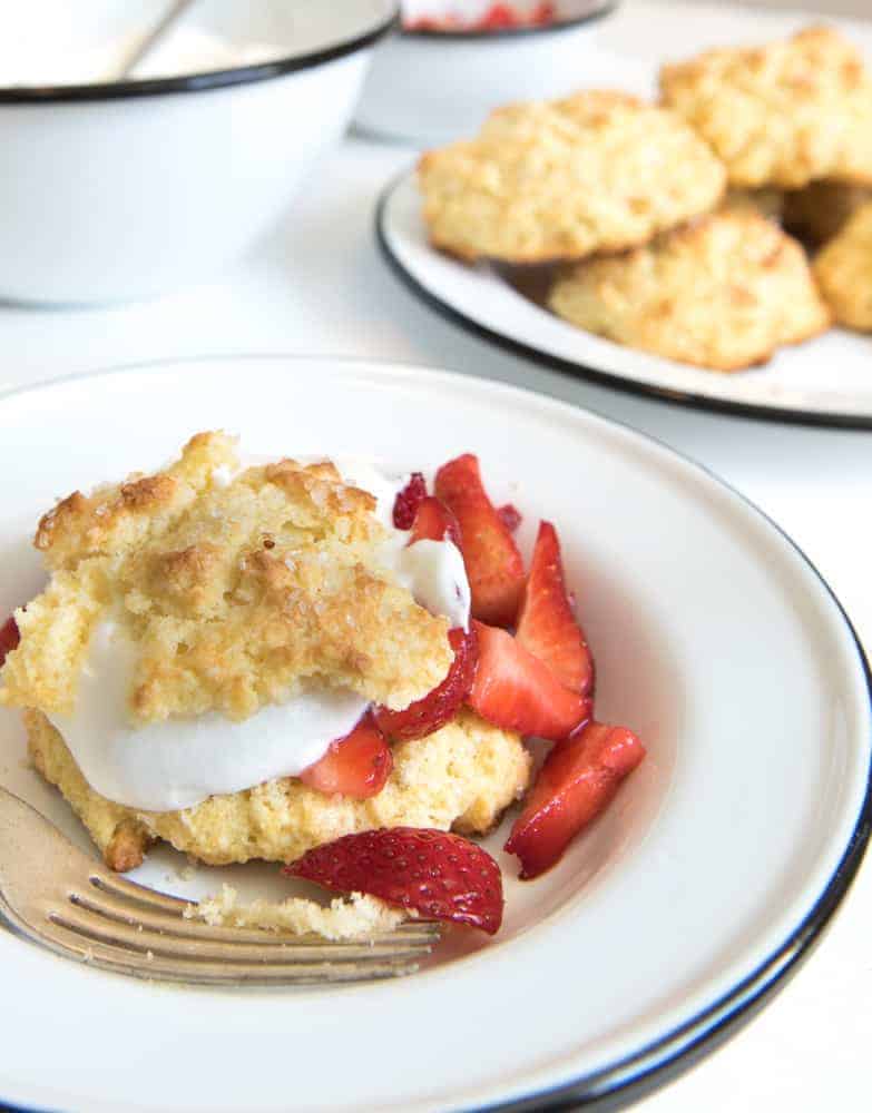 A plate with the best strawberry shortcake features a layer of sliced strawberries and whipped cream between crumbly biscuit pieces. A fork rests beside it, inviting a taste, while more biscuits tempt in the background.