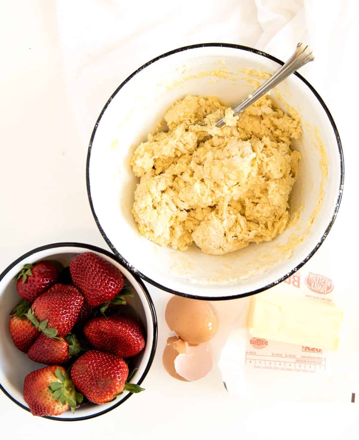 A white bowl with shortcake dough mixed up beside a bowl of red strawberries, eggs and butter. 