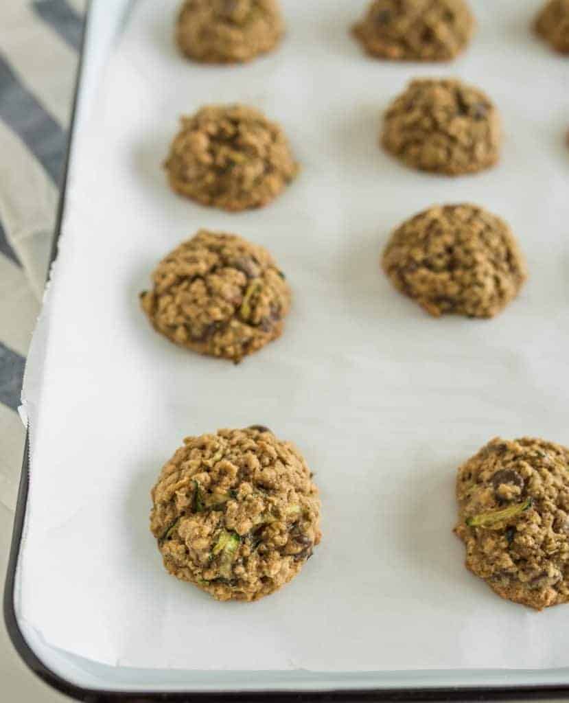 A baking tray lined with parchment paper holds several freshly baked, round cookies. Nestled beside the cookies are taco zucchini boats, offering a savory balance to the sweet treats. The cookies are spaced evenly and look slightly textured, suggesting ingredients like oats or nuts.