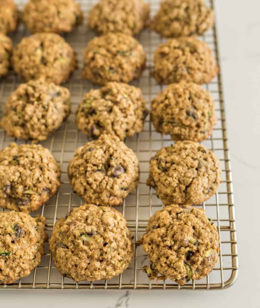 A cooling rack filled with freshly baked oatmeal cookies rests next to a tray of colorful taco zucchini boats. The cookies, round and golden-brown, are studded with chunks that hint at chocolate chips or nuts. They sit invitingly on a wire rack atop a pristine white surface.