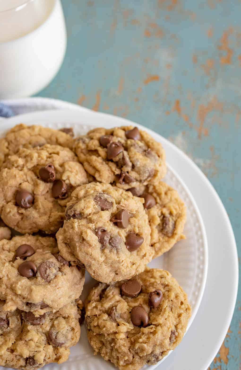 chocolate chip cookies piled on white plate with glass of white milk behind.