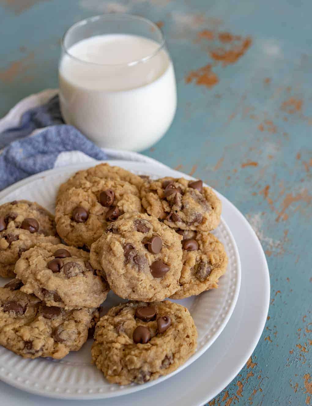 A plate of chocolate chip cookies sits on a rustic blue surface, accompanied by a glass of milk. A folded blue cloth is partially visible beside the plate.