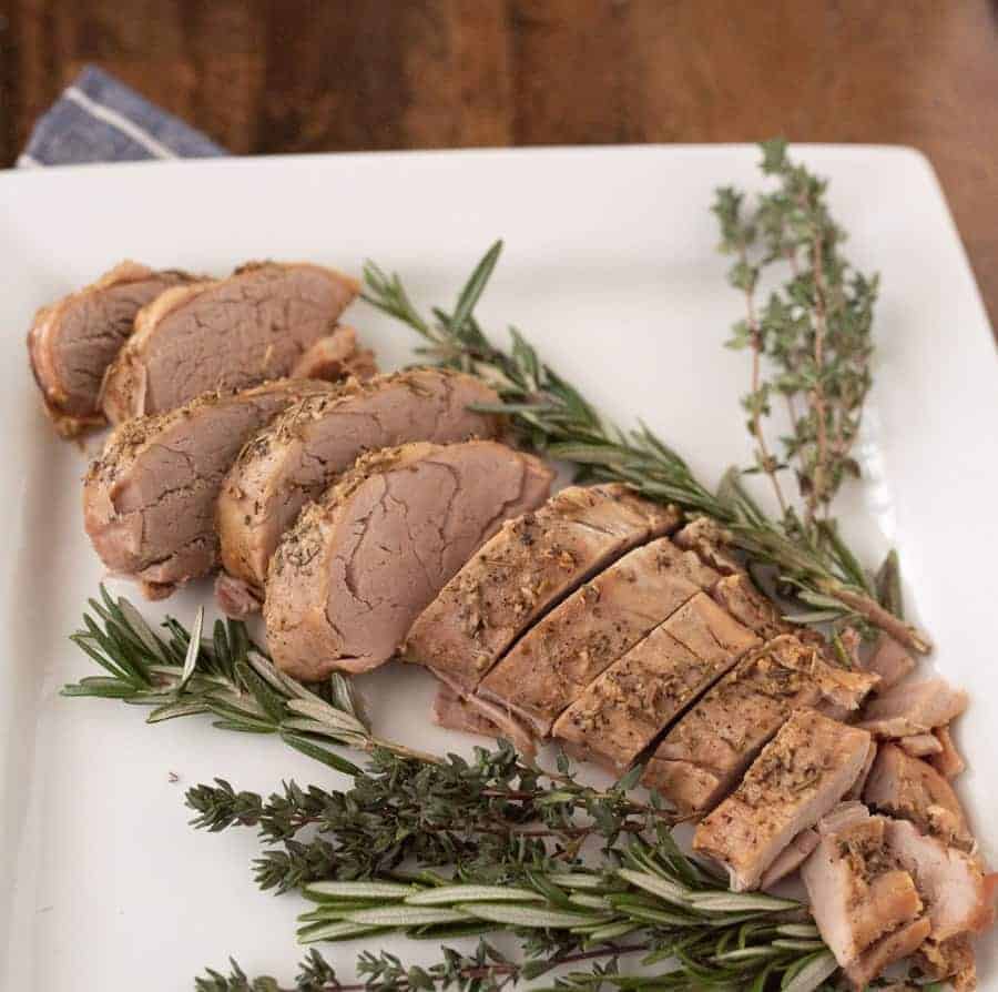 A plate of crock pot cooked pork loin, sliced, with fresh herbs around it. 