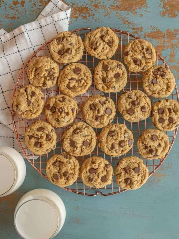 A cooling rack of peanut butter oatmeal and chocolate chip cookies rests on a rustic teal surface, accompanied by two glasses of milk. A checkered cloth is partially visible in the corner.