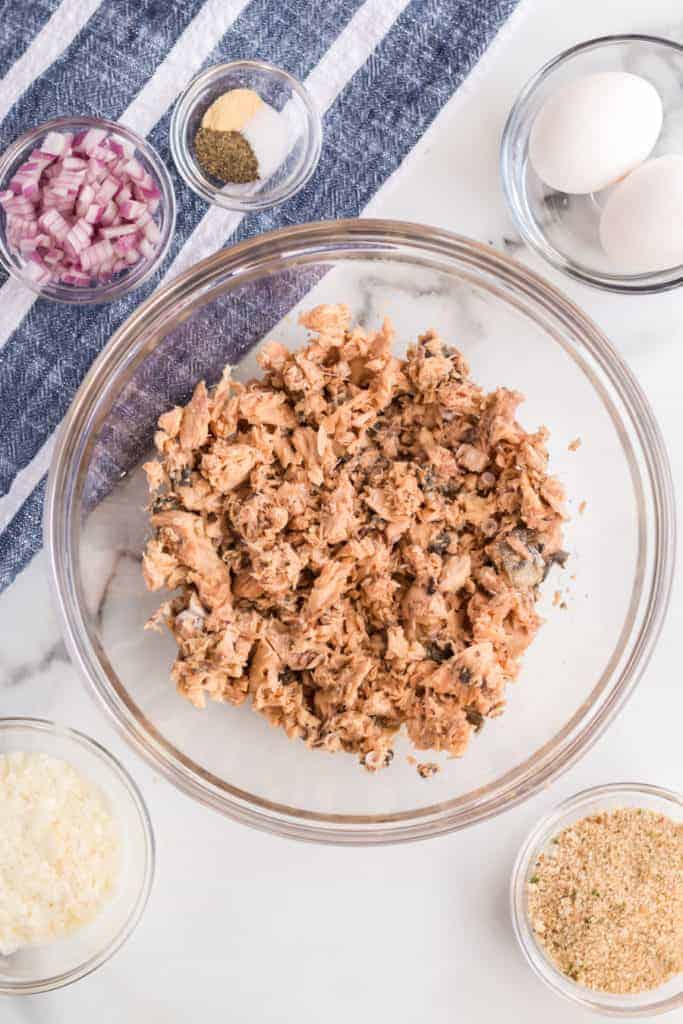 Top view of a cooking preparation scene featuring a glass bowl with shredded salmon at the center. Surrounding it are small bowls with chopped red onion, seasonings, breadcrumbs, grated cheese, and two whole eggs on a striped cloth.