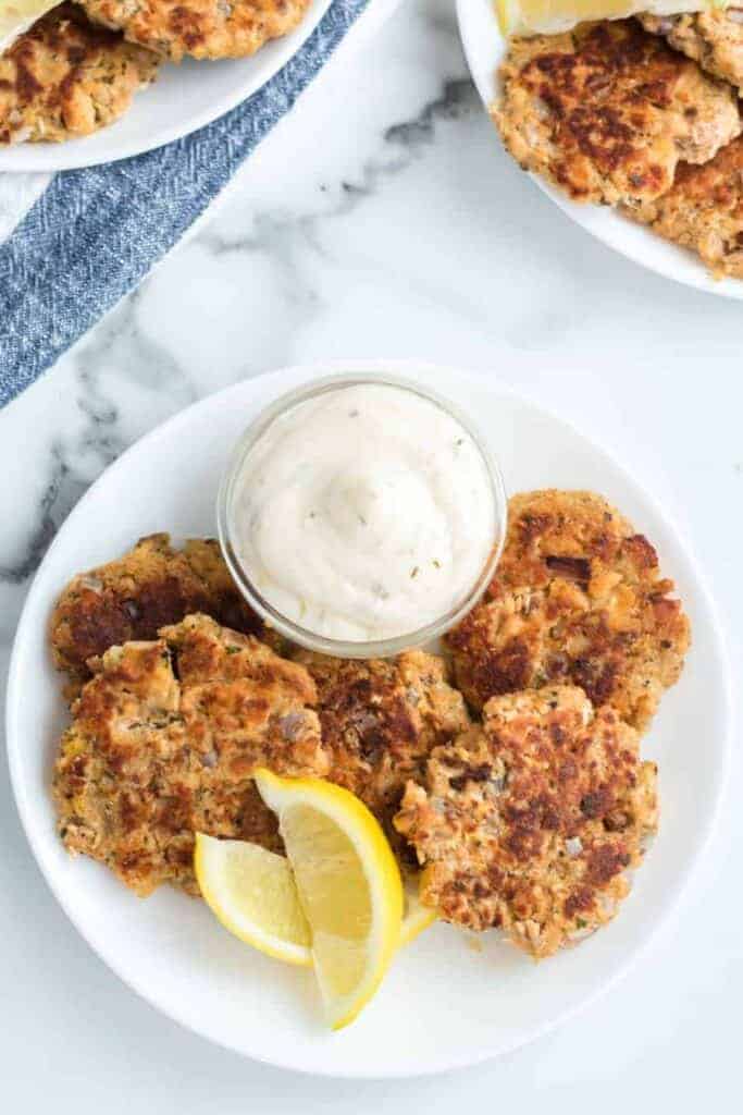 A white plate showcases four pan-fried salmon cakes, reminiscent of an old-fashioned salmon patties recipe, accompanied by a lemon wedge and a small glass bowl of creamy sauce. In the background, more salmon cakes are neatly arranged on the marble surface.