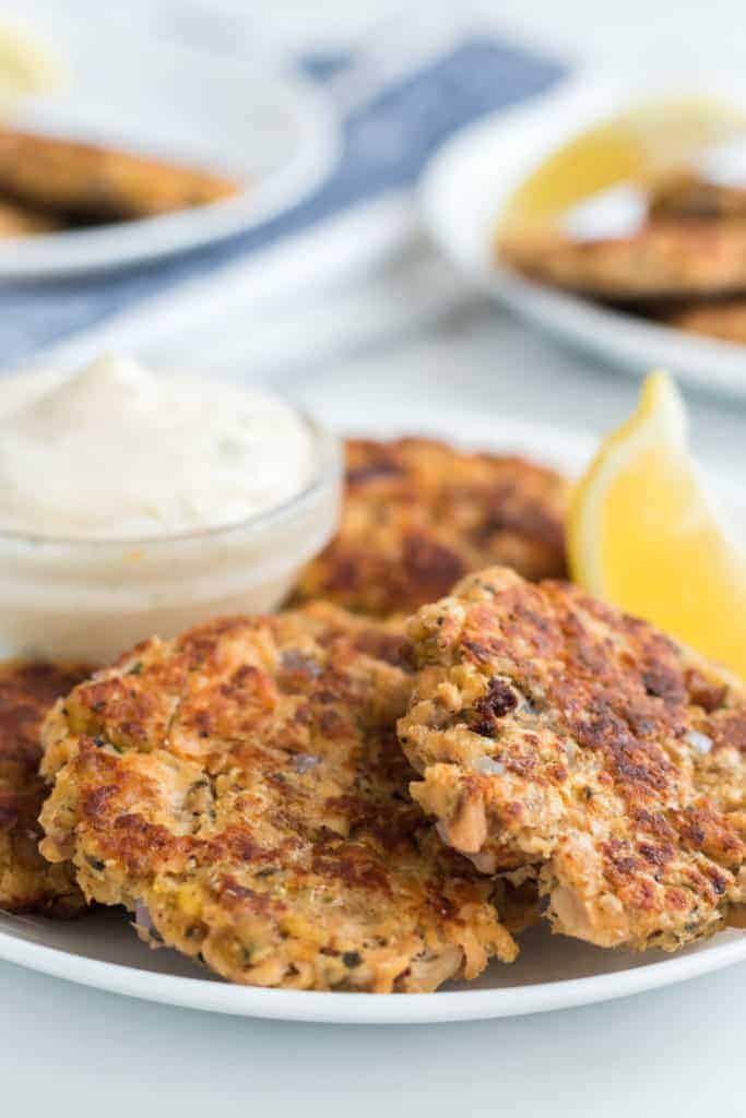 A white plate showcases golden brown salmon patties alongside a small glass dish of creamy sauce. A lemon wedge sits invitingly on the side. In the softly blurred background, another plate holds even more delectable salmon patties.