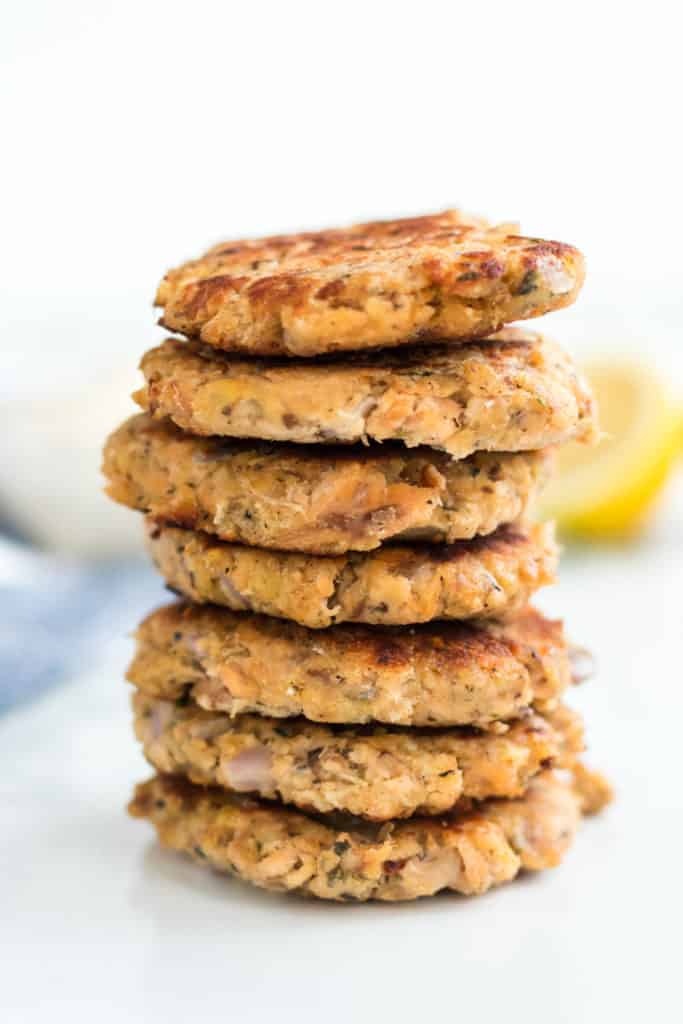 A stack of six golden-brown salmon patties, crafted using an old fashioned salmon patties recipe, rests on a white surface with a blurred lemon wedge in the background.