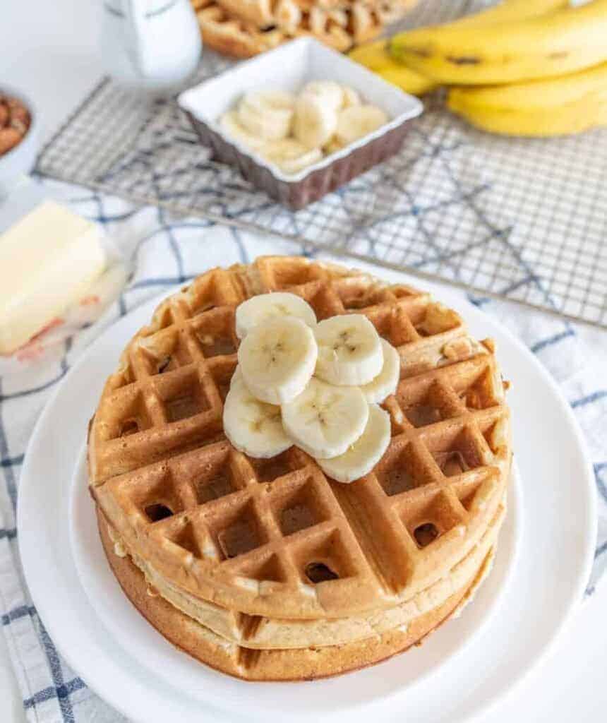 A stack of banana waffles topped with banana slices sits on a white plate. Nearby, theres a block of butter, an extra bowl of banana slices, a bunch of whole bananas, and a milk jug on a plaid tablecloth.