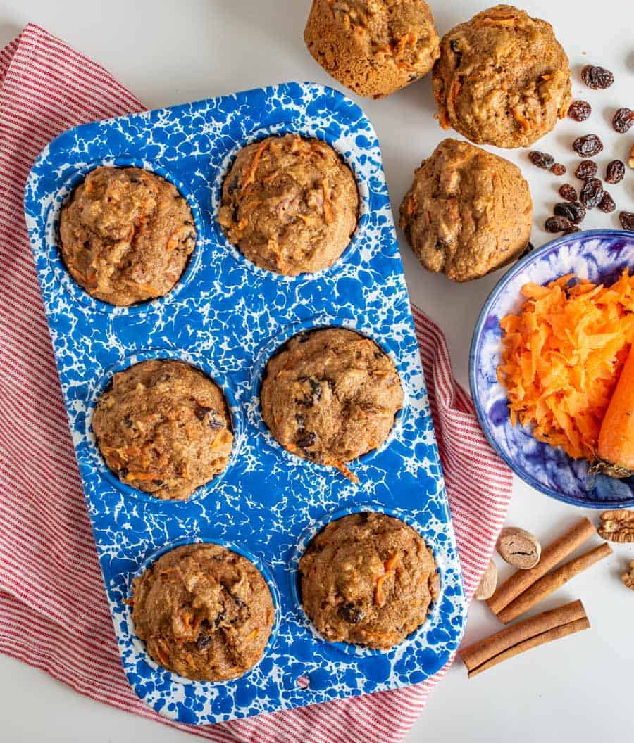 muffins in a blue and white muffin pan with small bowl of shredded carrot, cinnamon sticks, raisins, on a red and white striped towel.