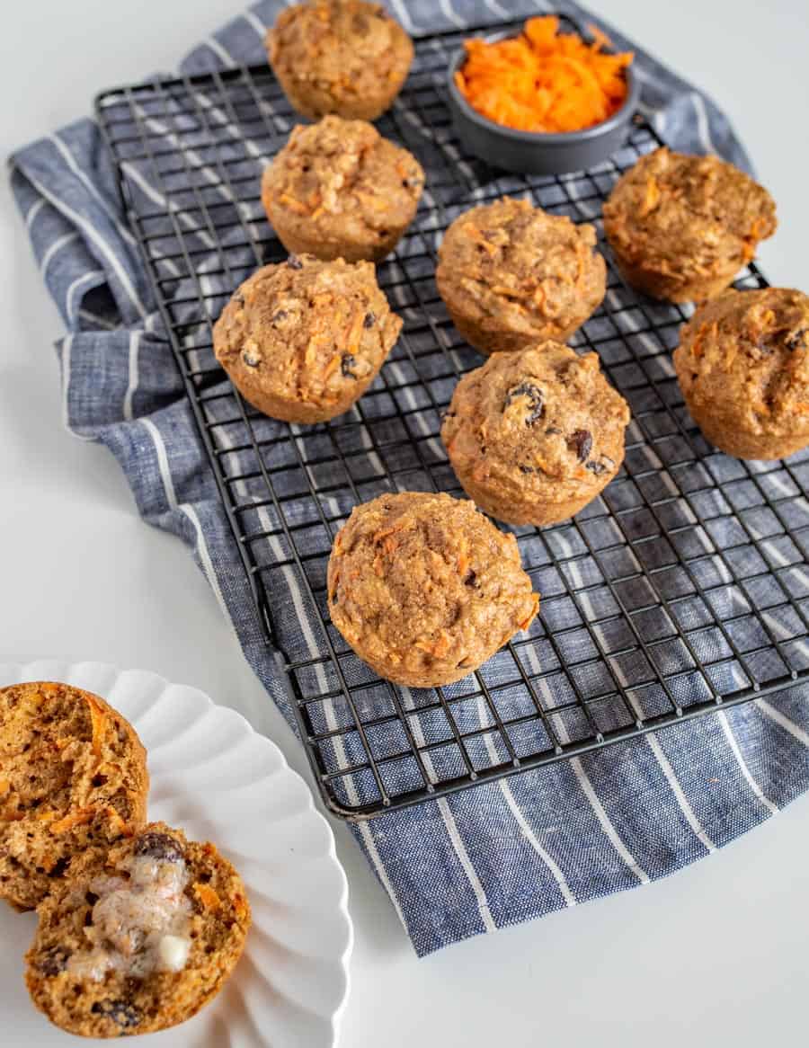 muffins on a metal rack with carrot pieces and raising showing through top with small bowl of shredded carrot on a blue and white striped towel.