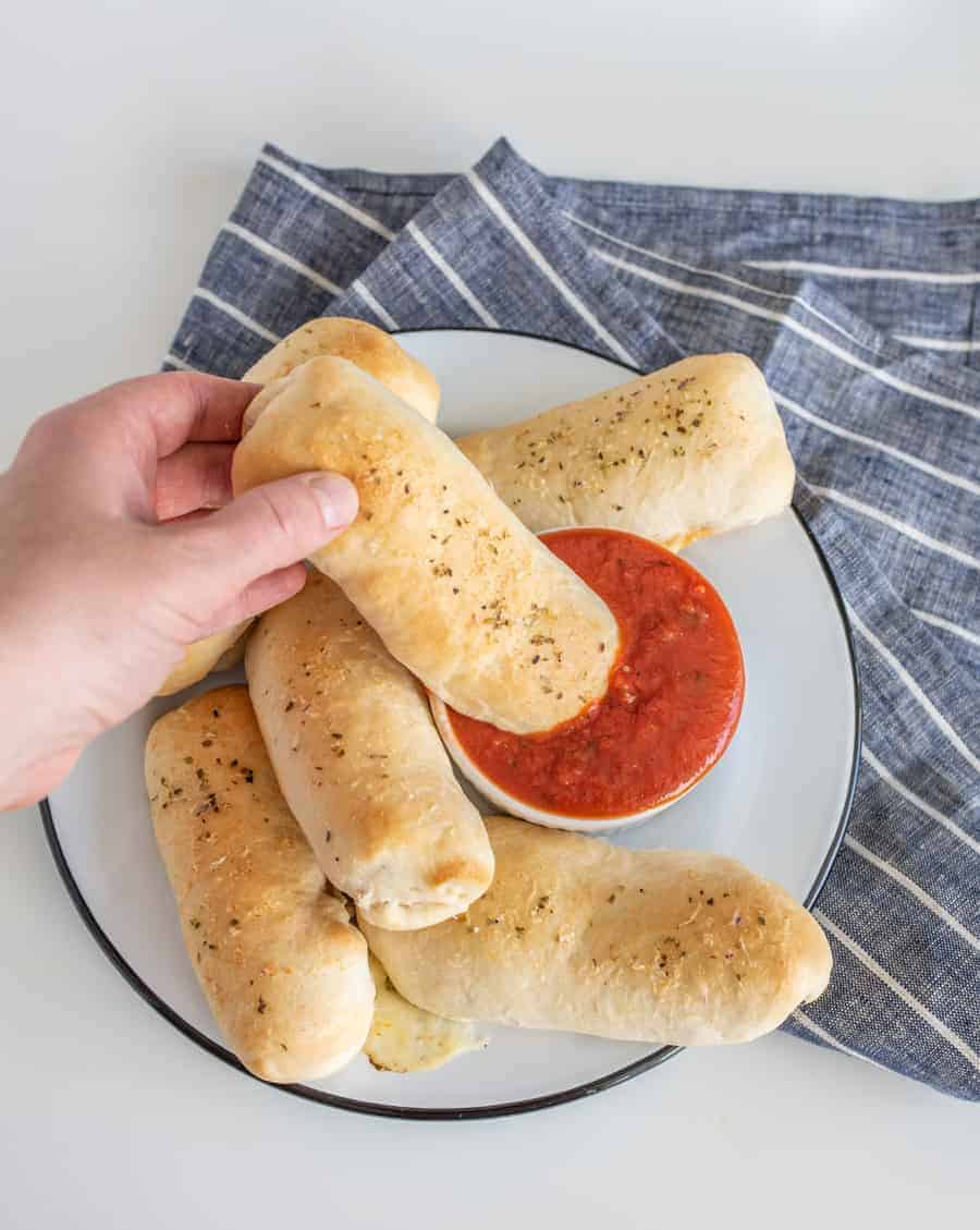 pizza sticks on a white plate with a small bowl of red marinara sauce on a blue and white striped towel with hand dipping one stick into sauce.