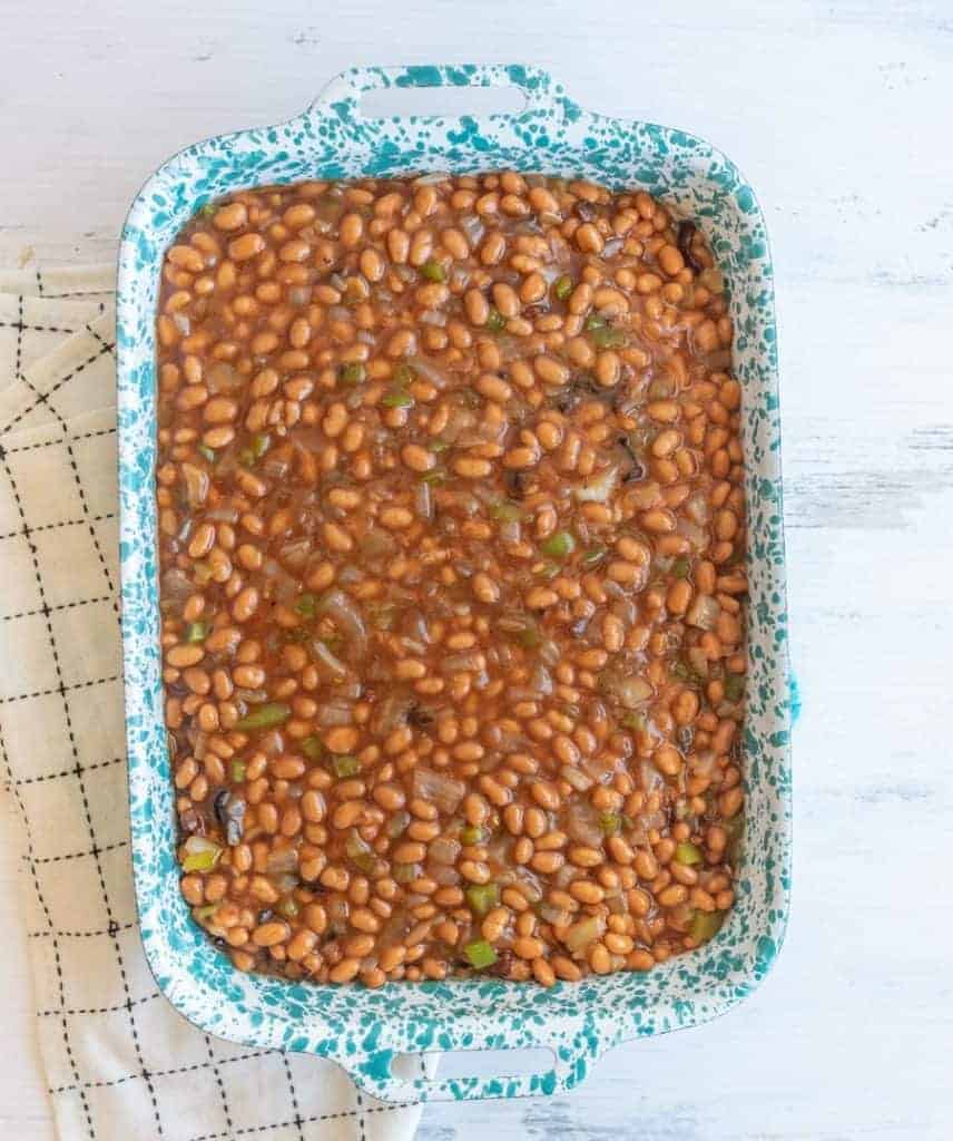 A blue speckled baking dish brims with savory baked beans, dotted with pieces of green pepper. Beside it, a white napkin with a black grid pattern rests on the crisp, white wooden surface.