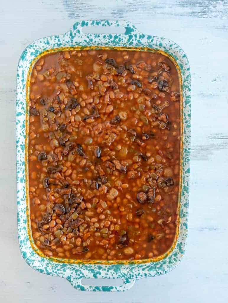 A blue speckled baking dish brimming with savory baked beans, crowned with chunks of crispy bacon. The beans are enveloped in a rich, dark sauce, creating a mouthwatering display as it rests on a white textured surface.