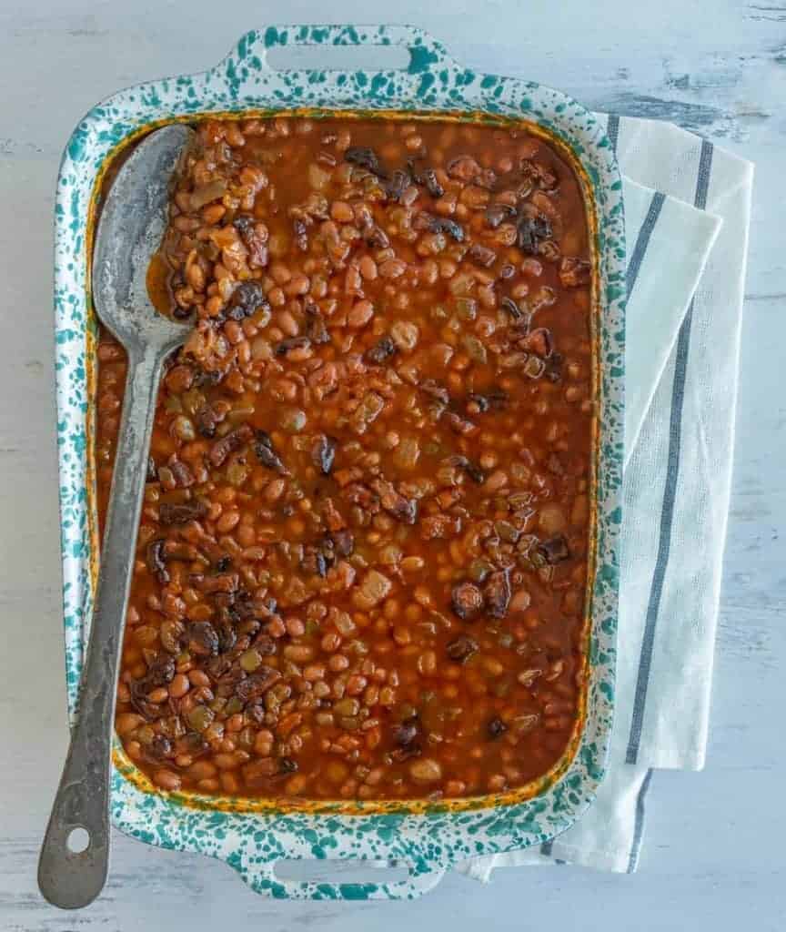 A baking dish brimming with savory baked beans in a rich, dark sauce sits invitingly. A large serving spoon rests on top, ready to serve. The dish is elegantly placed on a white wooden surface, complemented by a white and gray striped cloth underneath.