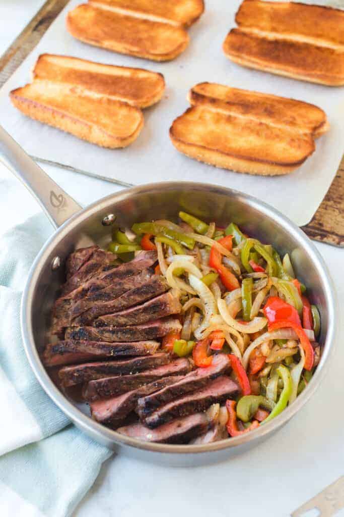 a pot of steak and veggies for a philly cheesesteak next to grilled buns