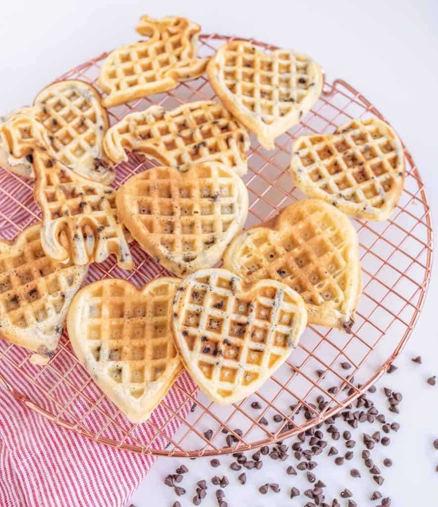 Heart-shaped and animal-shaped waffles rest on a round cooling rack atop a red-striped cloth, with small chocolate chips playfully scattered around. Imagine pairing these delightful treats with a scoop of your favorite ice cream for an indulgent treat. The background is a light, neutral color.