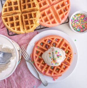 Three colorful waffles are on display, with one on a plate topped with a scoop of creamy ice cream and rainbow sprinkles. A bowl of delicious ice cream, a dish of sprinkles, and a red-striped cloth enhance the scene—a perfect pairing of waffles and ice cream.