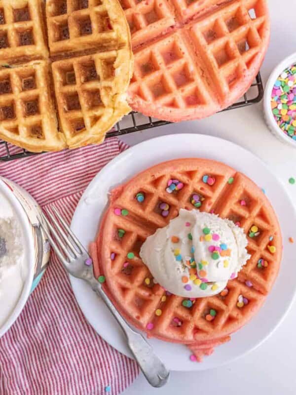 Three colorful waffles are on display, with one on a plate topped with a scoop of creamy ice cream and rainbow sprinkles. A bowl of delicious ice cream, a dish of sprinkles, and a red-striped cloth enhance the scene—a perfect pairing of waffles and ice cream.