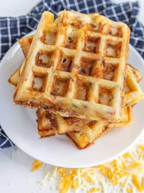 A stack of golden brown, square ham and cheese waffles sits on a white plate, resting on a blue and white checkered cloth. Shredded cheese is scattered around, hinting at the delicious ham and cheese tucked inside these savory waffles.