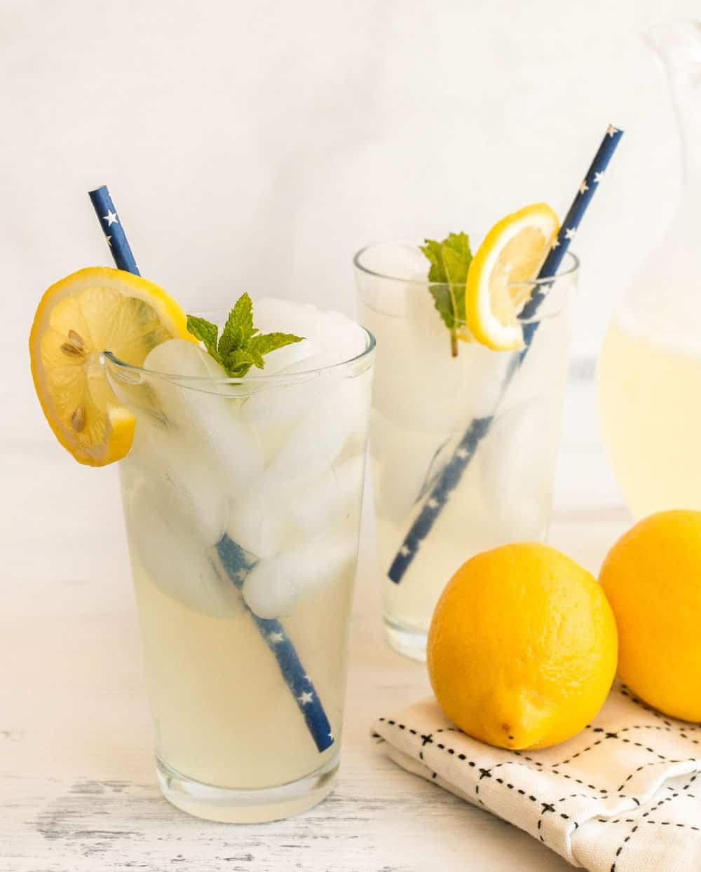 two glasses of lemonade with blue straws, ice, and a lemon and mint garnish