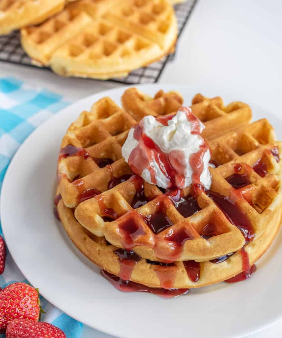 Stack of waffles on white plate with cream and red sauce on top and strawberries by plate.