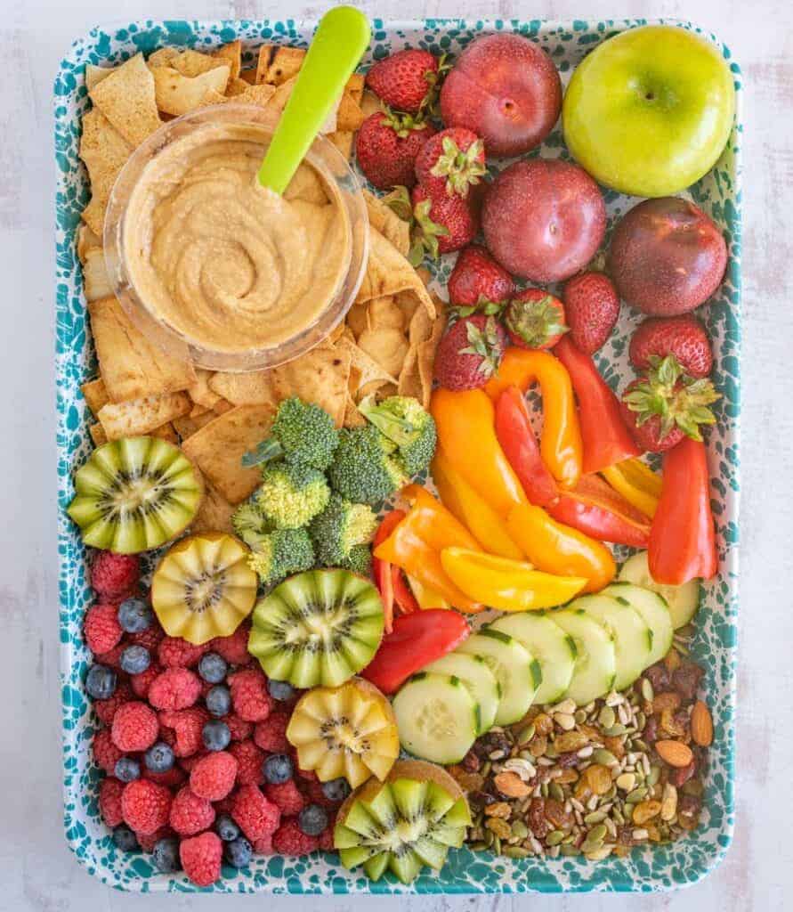 top view after school snack tray on a blue speckled sheet with mixed fruits, veggies, nuts, hummus and pita chips