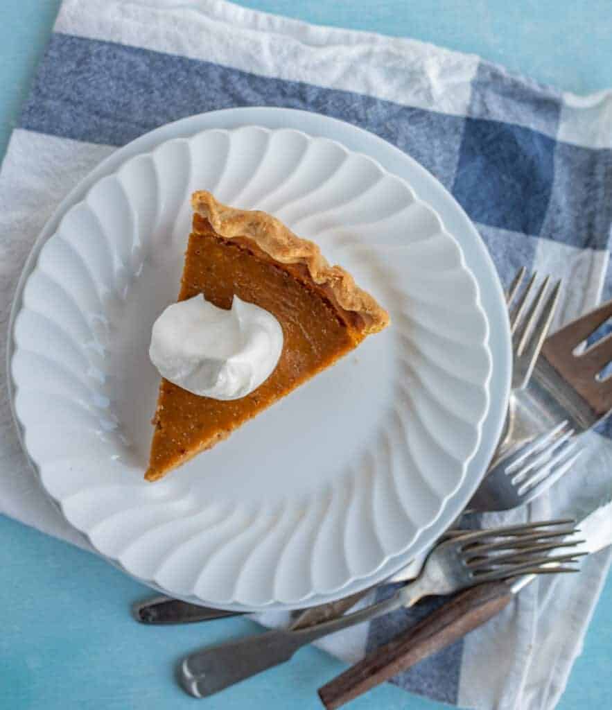 one slice of classic pumpkin pie with a dollop of cool whip on a white plate with forks around