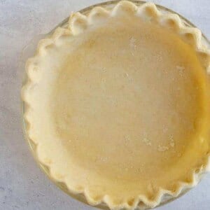 An all butter pie crust in a glass pie dish rests on a light gray surface. With a fluted edge, the unbaked crust displays a smooth, even surface tinged with a slightly golden hue. Nearby, a small piece of dough awaits its turn.