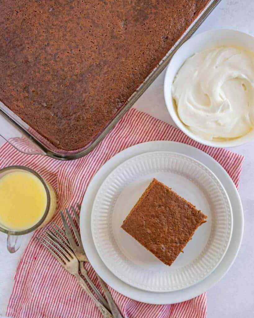 square of gingerbread cake taken out of the pan, onto a white plate and then iced and whipped cream on the side in white dishes - view from the top