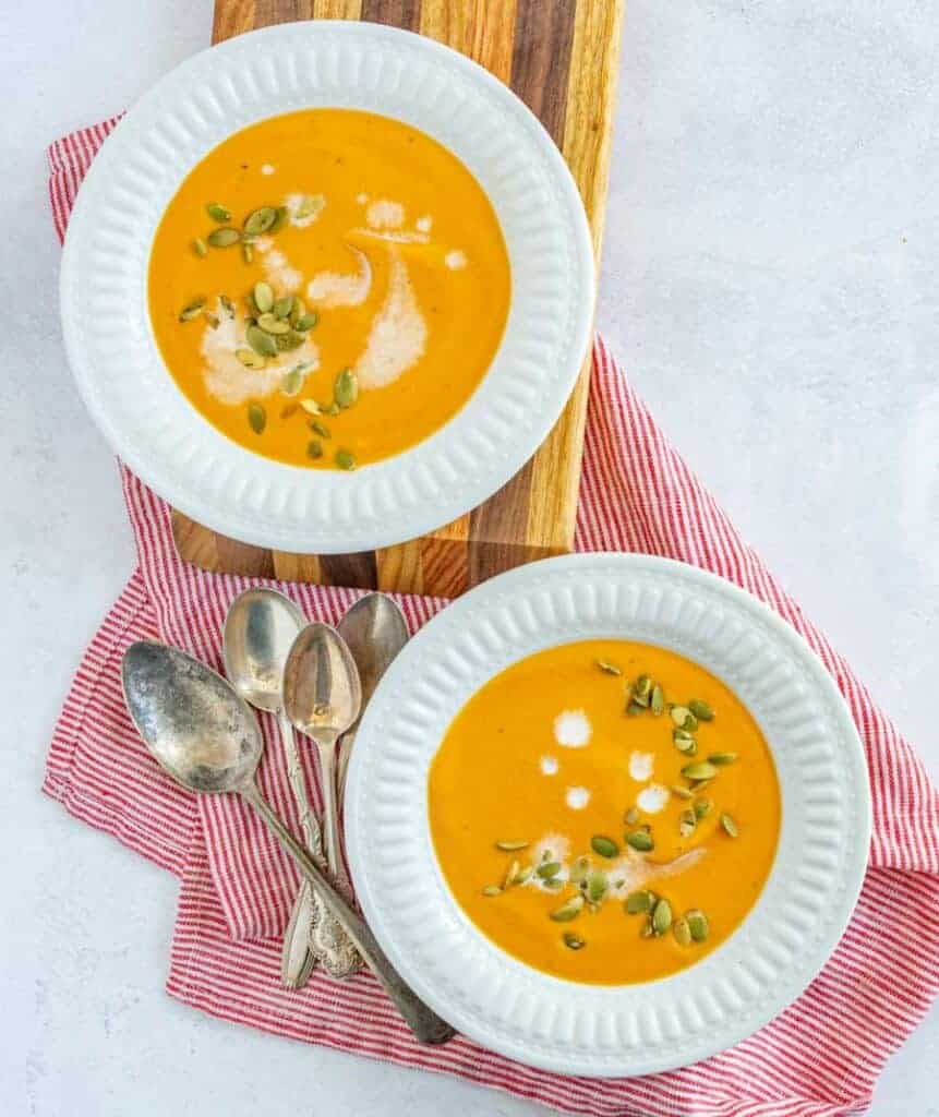 two white bowls of creamy carrot soup with creme fresh and chives on top with spoons next to it
