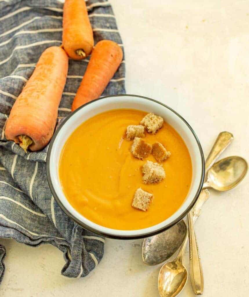 photo of a bowl of creamy carrot soup with a row of croutons in it a raw whole carrots in the background